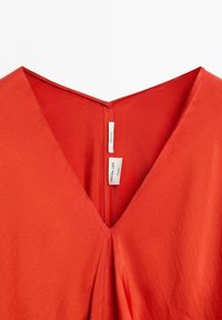 Rood katoenen kledingstuk met een V-hals. De stof heeft een gladde textuur en heeft een centrale plooi met een verborgen rits aan de achterkant.