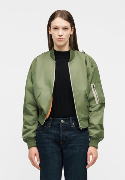 Femme portant une veste bomber verte par-dessus un pull noir et un jean bleu foncé, debout devant un fond blanc uni.