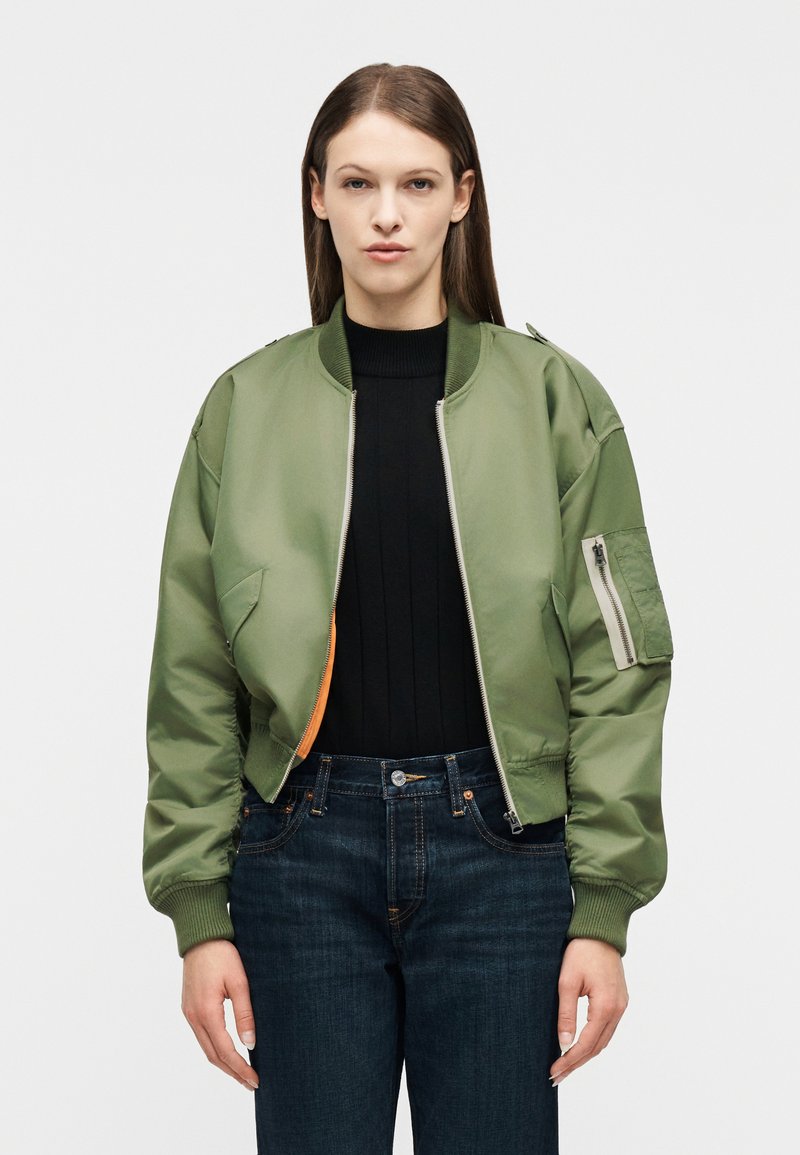 Γυναίκα που φοράει πράσινο μπουφάν τύπου bomber πάνω από μαύρο πουλόβερ και σκούρο μπλε τζιν, στέκεται μπροστά σε απλό λευκό φόντο.