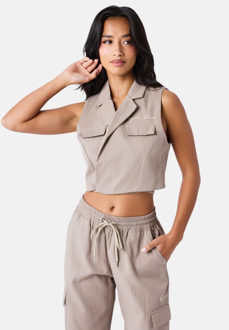 Blazer sans manches en tissu beige clair avec deux poches poitrine et des broderies subtiles. Associé à un pantalon à cordon de serrage assorti.