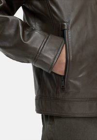 Bruine leren jas met een gladde textuur, een ritszak aan de zijkant en een getailleerde pasvorm. Zichtbare stiksels en detailwerking aan de mouwen.