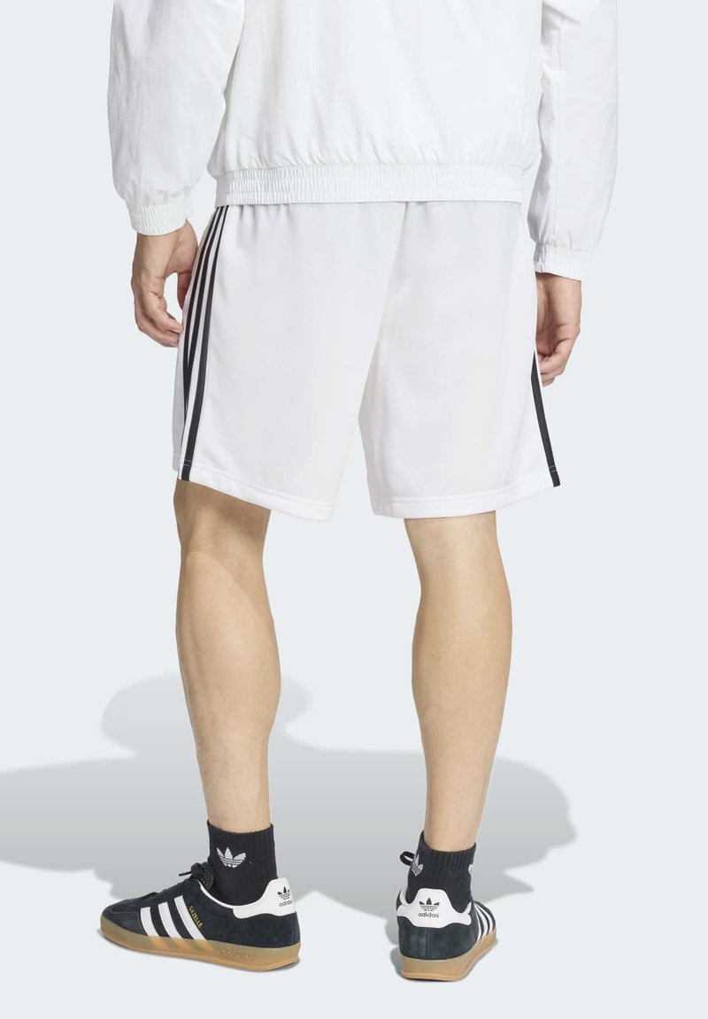 Persona che indossa pantaloncini sportivi bianchi con strisce nere, giacca bianca, calzini neri Adidas e scarpe da ginnastica nere Adidas con suole in gomma, vista da dietro.