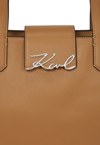Sac fourre-tout en cuir tan avec une fermeture à rabat magnétique, présentant un logo "Karl" en argent à l'avant. Texture lisse avec des coutures visibles.