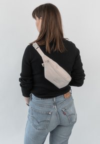 Sac bandoulière beige en matériau lisse, porté dans le dos. Dispose d'une fermeture éclair et d'un logo en relief. Associé à un sweater noir à côtes et un jeans bleu.