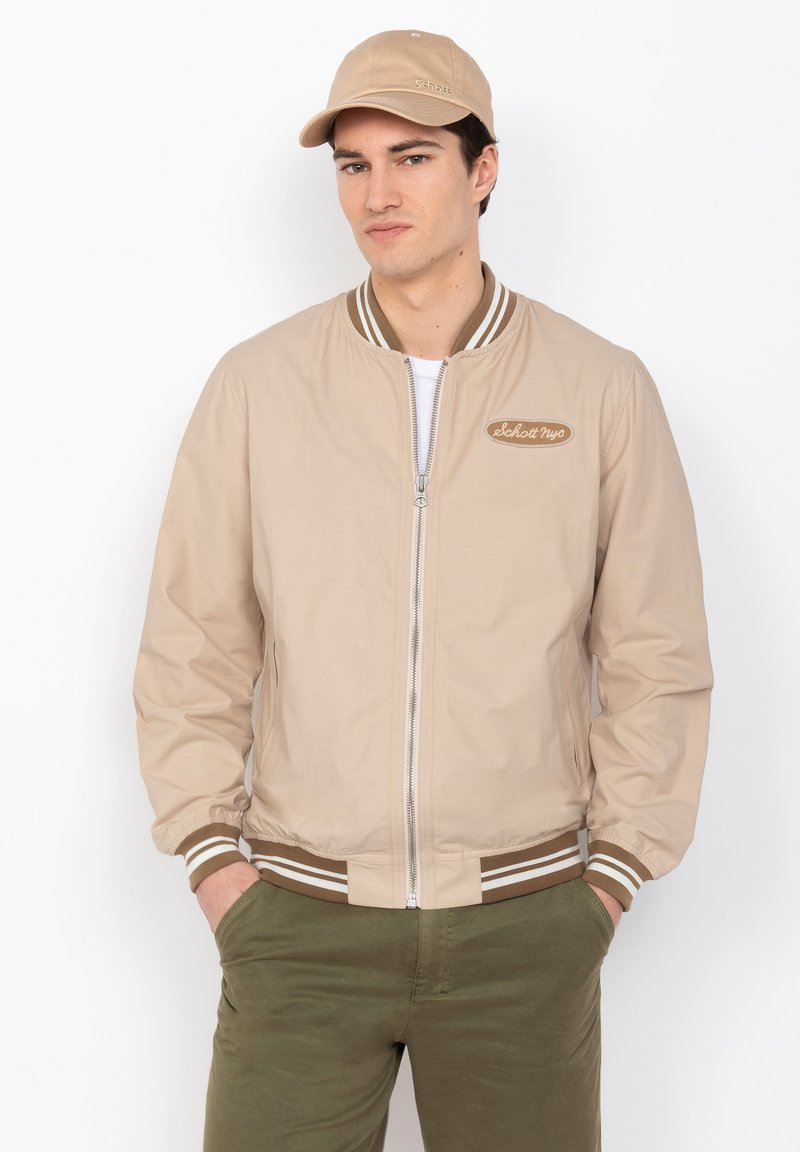 Beige Bomberjacke mit einem Reißverschluss vorne, gestreiften Rippbündchen und -saum sowie einem gestickten Logo auf der Brust. Getragen mit einer passenden Mütze und grünen Hosen.
