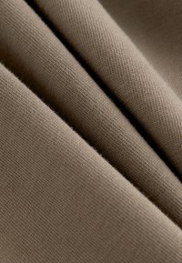 Brauner Strickstoff mit weicher Textur, bestehend aus mehreren gefalteten Lagen. Die Oberfläche zeigt ein feines, gleichmäßiges Muster mit leichtem Glanz.