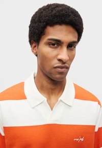 Polo às riscas laranja e branco com gola. Tecido macio e texturizado com um pequeno logótipo bordado no peito.
