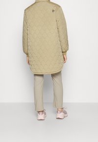 Beżowa pikowana długa kurtka z kołnierzem stójką, elastycznymi mankietami i małym czarnym logo na plecach. W zestawie z jasnymi spodniami i różowymi trampkami.