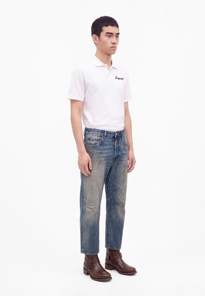 Jeune homme portant un polo blanc, un jean bleu délavé et des bottines en cuir marron, debout devant un fond blanc uni.