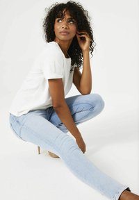 Camiseta blanca de manga corta y jeans ajustados azul claro; modelo en cuclillas con cabello rizado, mostrando un atuendo casual contra un fondo liso.