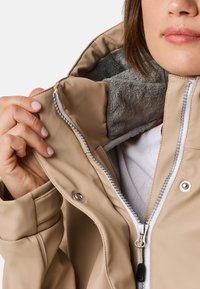 Veste imperméable beige avec une douce doublure en polaire grise, fermeture éclair à l'avant et boutons-pression, tenue ouverte pour montrer les détails intérieurs.
