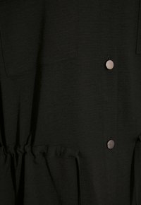 Zwarte kledingstuk met twee borstzakken, zilveren drukknoopjes en een elastische taille. Textuurstof met een comfortabele pasvorm en op maat gemaakte naden.