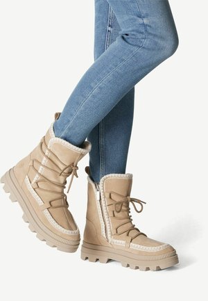 Lysebrune vinterstøvler med tykke såler, snørelukning foran, lynlåse i siden og hvid kant, båret med blå tætsiddende jeans.