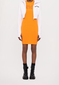 Orange figursydd klänning kombinerad med en kort vit hoodie med en logotyp. Svarta ankelboots med snörning och spännen fullbordar looken.