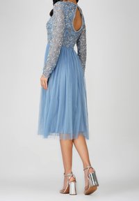 Vestido azul de encaje con mangas largas y transparentes, y un escote en forma de lágrima en la espalda. Falda semitransparente. Combinado con sandalias de tacón bloque metálicas plateadas.