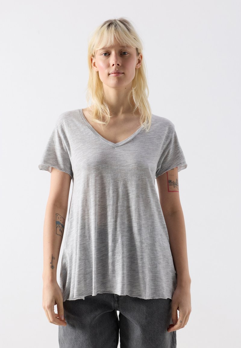 Blonde sieviete ar tetovējumiem uz rokām, valkājot vaļīgu pelēku V-veida kakla T-kreklu un tumšas bikses, stāvot pret vienkāršu baltu fonu.