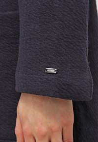 Marlowá texturovaná látková rukáv s stříbrným obdélníkovým štítkem s nápisem "HILFIGER" připevněným poblíž manžety. V materiálu je viditelný jemný vzor.