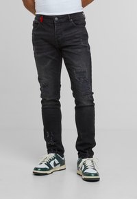 Man in versleten zwarte jeans met turquoise en witte sneakers, staand op een effen lichtgrijze achtergrond.