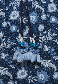 Mörkblåt blommönstrad tyg med vita och ljusblå blommor, försett med flätade band dekorerade med blå och svarta pärlor samt ljusblå fransar.