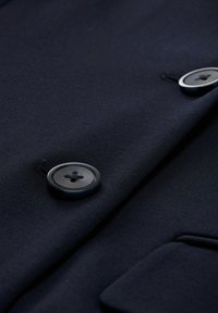 Material bleumarin, prim-plan al unei jachete cu un buton rotund proeminent cu patru găuri, textură netedă și detaliu de buzunar.