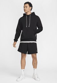 Felpa nera con tasca frontale, cordini bianchi, abbinata a pantaloni corti neri. Logo del marchio ben visibile sui pantaloni corti e sneakers bianche.
