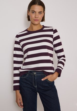 Femme aux cheveux courts et bruns porte une chemise à manches longues avec de larges rayures horizontales blanches et violet foncé et un jean bleu foncé.