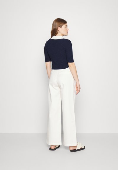 Top de malha de manga curta azul-marinho com colarinho branco, combinado com calças brancas de cintura alta e sandálias pretas rasas. Fundo plain.