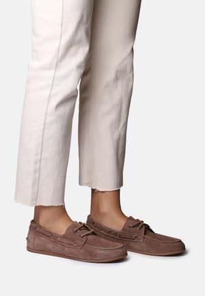 Dolne partie nóg w offwhite’owych, skróconych spodniach i brązowych mokasynach zamszowych ze sznurowadłami, stojące na białym tle.