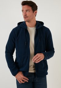 Felpa con zip di colore blu navy, con cappuccio regolabile, tasche a marsupio e finitura testurizzata. Indossata sopra una maglietta di colore chiaro.
