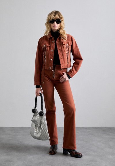 Jachetă din denim de culoare ruginie cu capse argintii, asortată cu pantaloni evazați. Modelul poartă o geantă din plasă metalică și ochelari de soare negri.