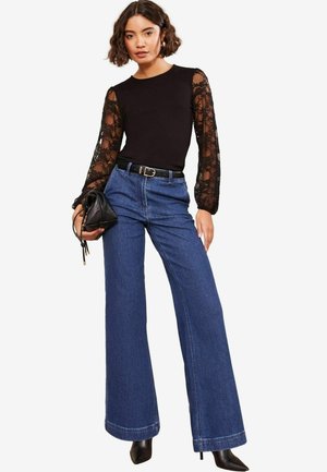 Haut noir avec des manches en dentelle, associé à un jean en denim bleu taille haute et large, et un sac à main noir. Des talons aiguilles complètent la tenue.