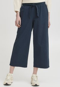 Pantalons larges bleu marine avec une taille à cordon, fabriqués en tissu doux, présentant des poches latérales et une coupe décontractée, associés à des baskets légères.