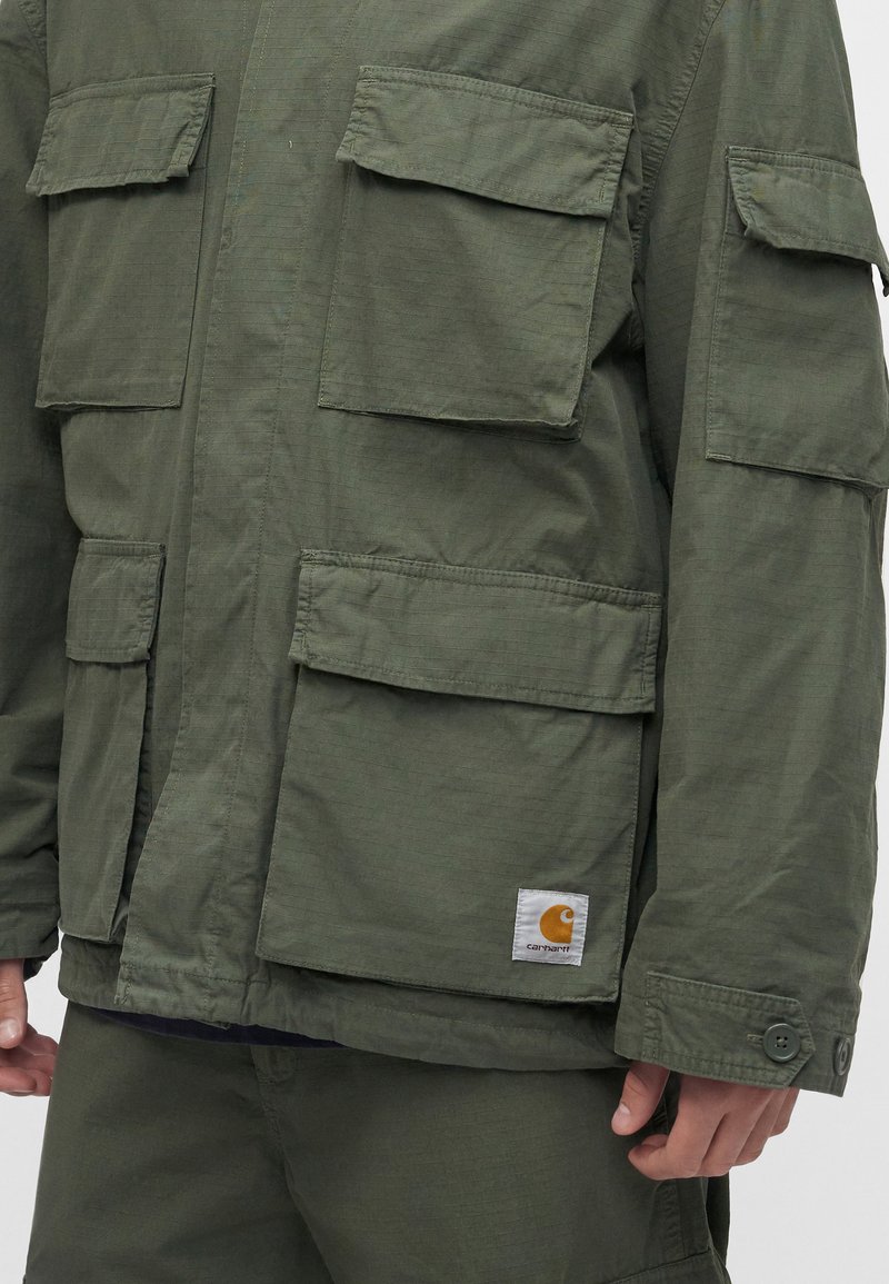 Hombre que lleva una chaqueta verde oliva con múltiples bolsillos con solapa y pantalones a juego, mostrando un pequeño logo rectangular de la marca en el bolsillo.