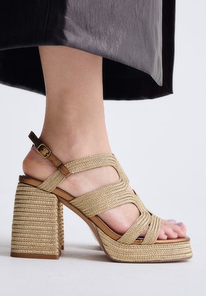 El calzado muestra una sandalia tejida color marrón claro con tacón bloque y pulsera en el tobillo, llevada por una persona con dedos visibles, combinada con un dobladillo de falda negro.