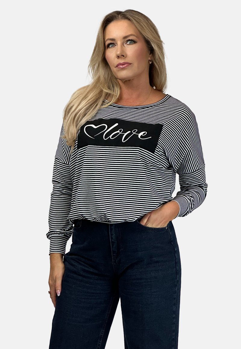Kobieta z długimi blond włosami, ubrana w czarno-białą pasiasta bluzkę z długim rękawem, na której znajduje się napis "love" oraz serce na klatce piersiowej, oraz czarne spodnie, trzymająca rękę w kieszeni.