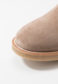 Beige mokkanahka kengät, joissa on pyöreä kärki, ommeltu yksityiskohta kantapään ympärillä ja luonnollisen värinen kumipohja. Sileä ja teksturoitu pinta.