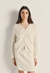 Lehký cardigan smetanové barvy s výstřihem do V, zapínáním na knoflíky a žebrovaným lemem; jemná textura, v kombinaci s bílou sukní.