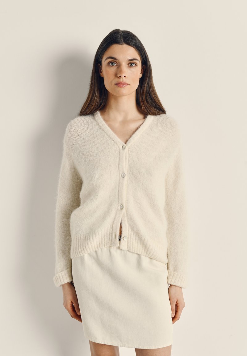 Lehký cardigan smetanové barvy s výstřihem do V, zapínáním na knoflíky a žebrovaným lemem; jemná textura, v kombinaci s bílou sukní.