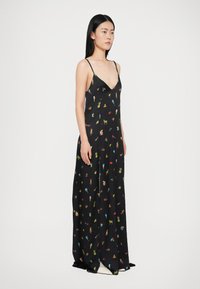 Frau mit langen schwarzen Haaren, die ein schwarzes Maxikleid mit bunten kleinen Drucken trägt, darunter Eiscreme, Ananas und Tiere, steht vor einem weißen Hintergrund.