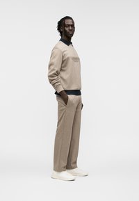 Sweat-shirt beige avec un motif brodé sur la poitrine, associé à un pantalon beige assorti. Le modèle porte des baskets blanches et un col bleu marine en dessous.