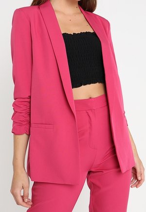 Femme portant un blazer rose vif à manches froncées et un pantalon assorti avec un crop top noir texturé, debout devant un fond blanc.