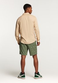 Maglietta beige chiaro a maniche lunghe, pantaloni corti verde con motivo a diamante e sneaker verde/nero. Vista posteriore semplice senza dettagli visibili.