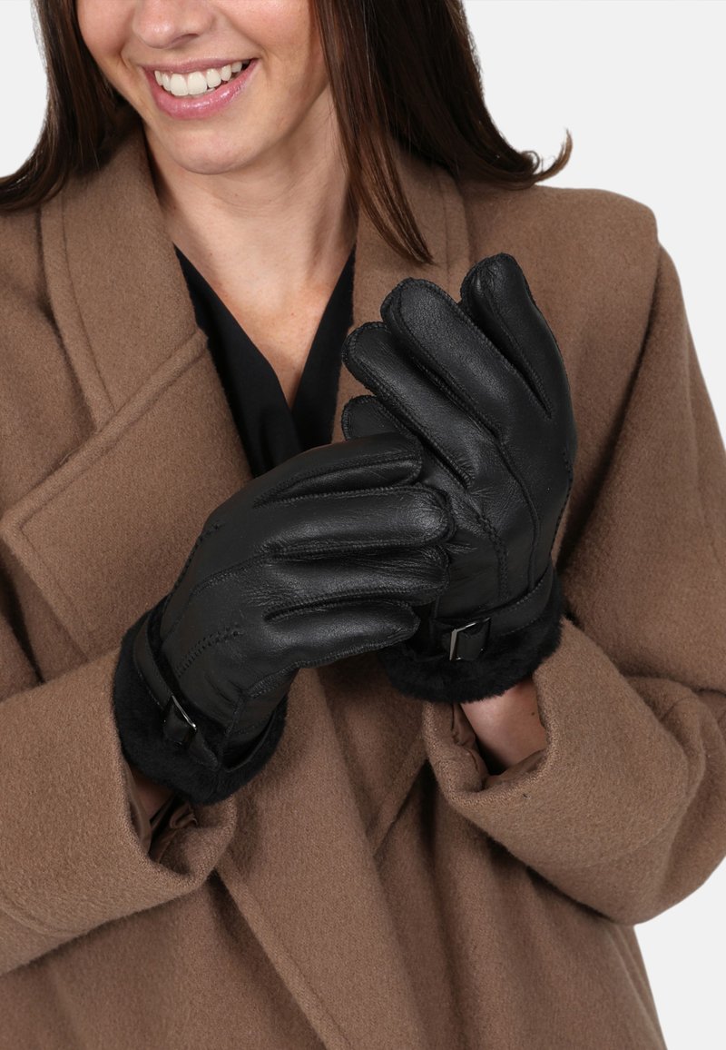 Black leather gloves with a soft fur lining and a wrist strap detail, worn by a person in a brown coat, showcasing a sleek design.