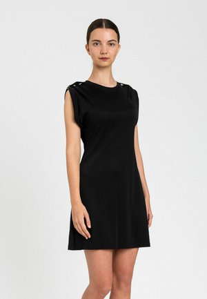 Donna con capelli scuri legati indietro che indossa un vestito nero senza maniche con dettagli a bottoni sulla spalla, in piedi davanti a uno sfondo bianco uniforme.