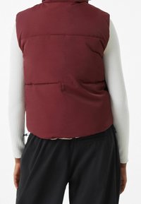 Bordowy kamizelka puchowa o gładkiej fakturze, pikowany design i wysoki kołnierz, noszona na białej koszuli z długim rękawem oraz zestawiona z czarnymi spodniami.