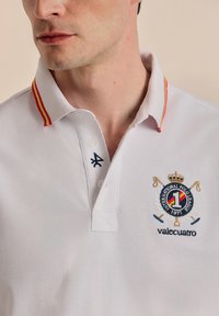 Camisa polo blanca con una tela texturizada, cuello a rayas rojas y amarillas, y un logo bordado que presenta una corona y unos palos de polo cruzados.