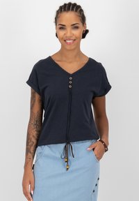 Top blu navy a V con maniche arrotolate, bottoni in legno decorativi e dettaglio con cordoncino in vita, abbinato a una gonna azzurro chiaro.