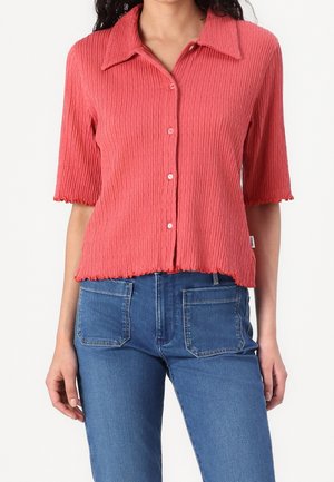 Kvinde iført en ribbet, knappet koralrosa kortærmet skjorte kombineret med blå højtaljede jeans med forlommer med lapper.