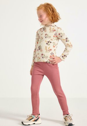 Ung barn med krulligt rött hår bär en blommig långärmad tröja, ribbade rosa leggings och mångfärgade sneakers, står med handen på höften.