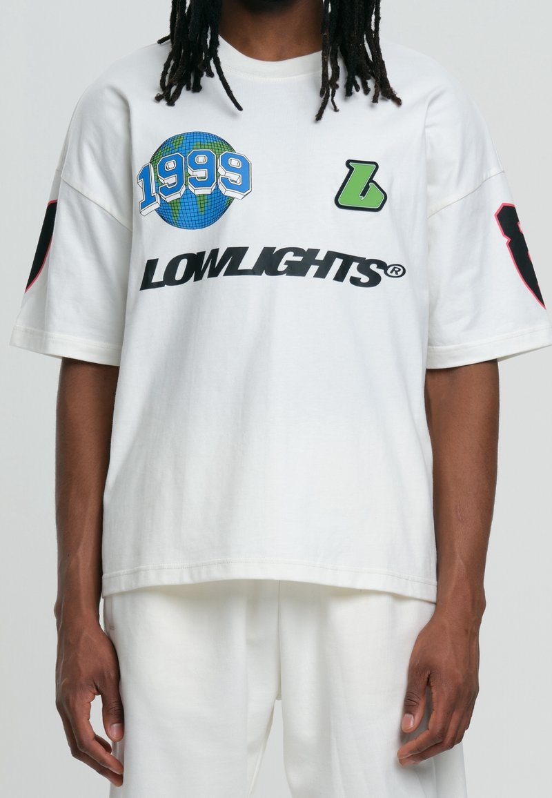 Homme portant un T-shirt blanc oversize avec des graphismes incluant un globe portant la mention "1999", un "L" vert et le texte "LOWLIGHTS" à l’avant.